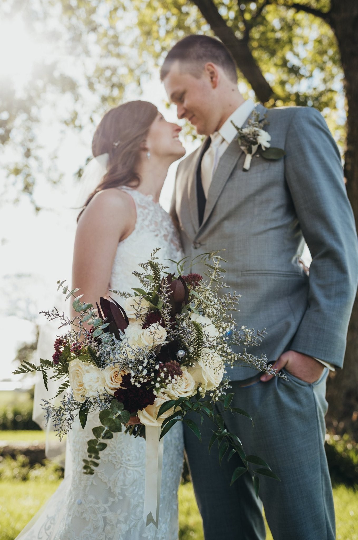 Newlywed couple is standing outside, under a large tree. Couple is smiling and looking ar each other. Bride is holding large bouquet loosely at her side.