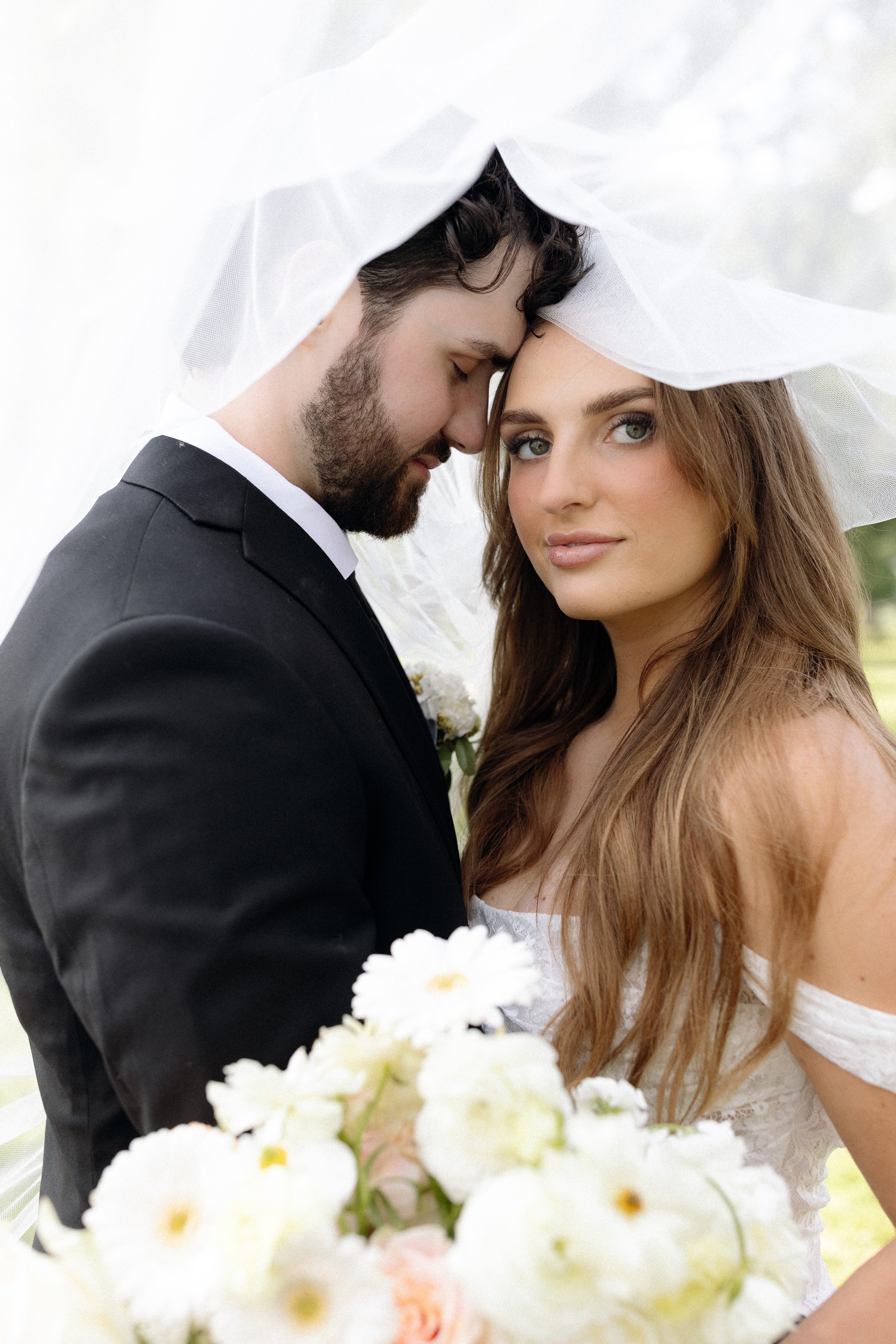 A newlywed couple standing outside. Groom is looking at the bride. Bride is looking at us, with bouquet in foreground, slightly blurry