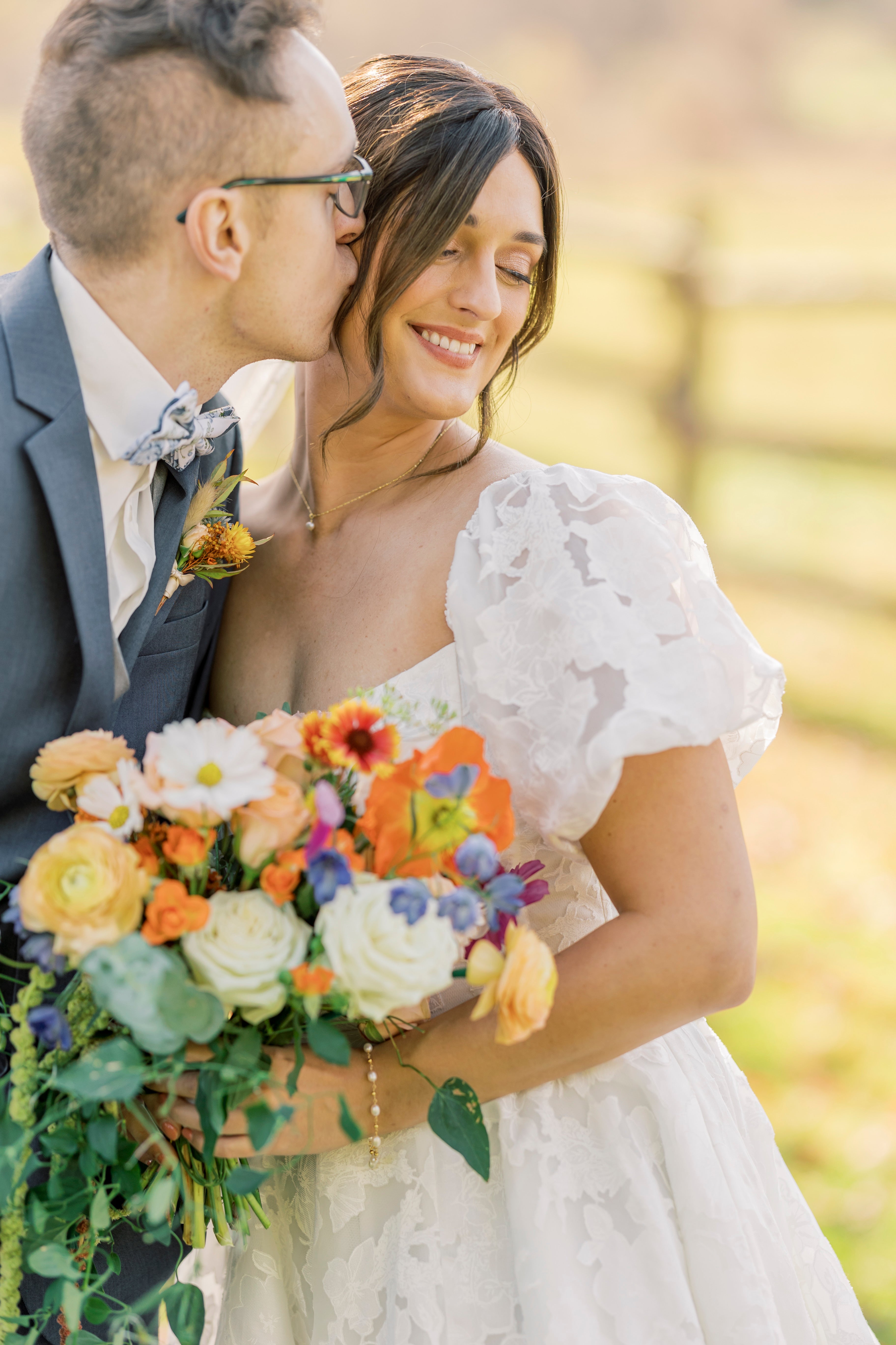 Newlywed couple is outside with a sunny field in the background. Groom is in a blue suit, kissing brides cheek. Bride is sming, looking down at large, colorful bouquet