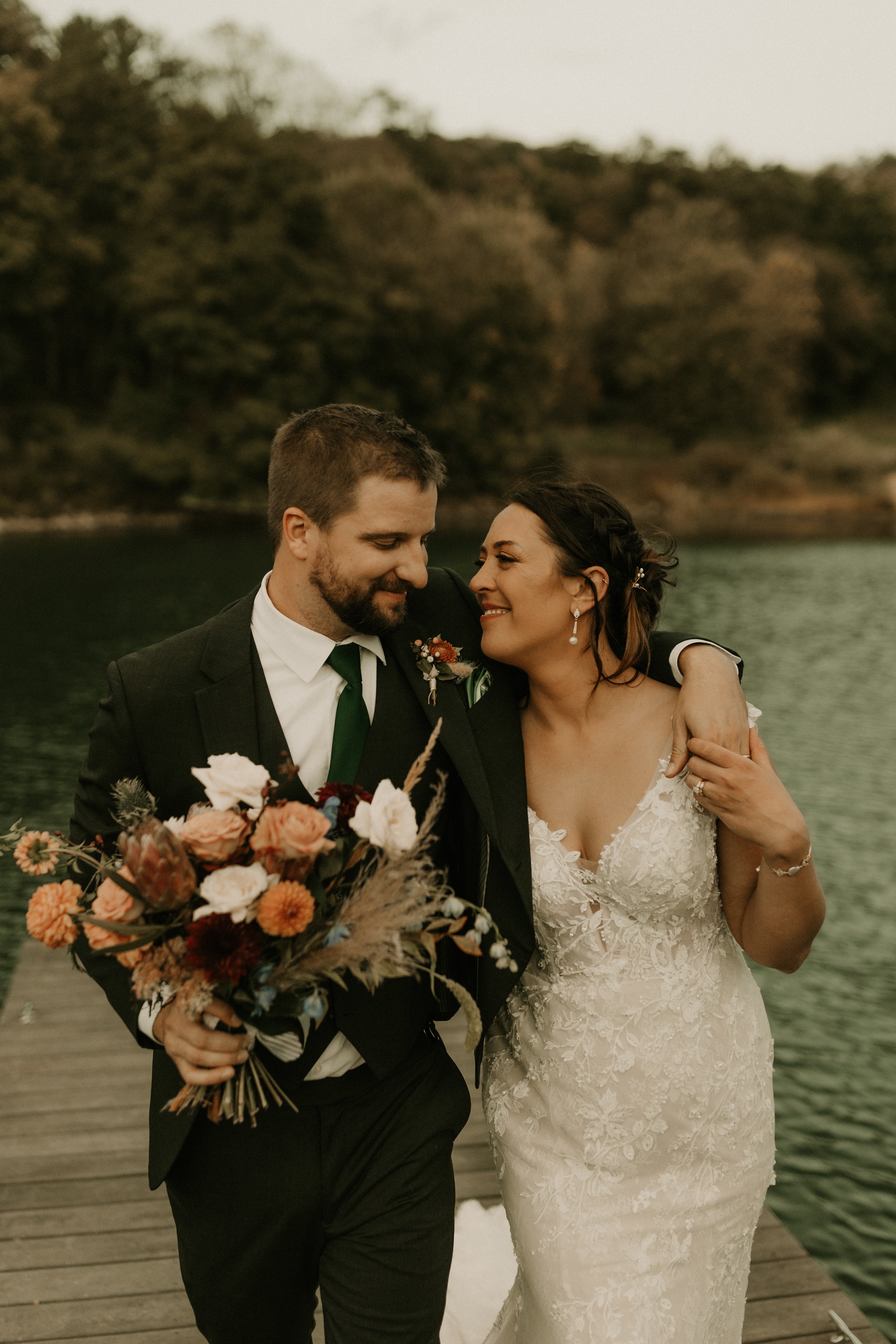 A smiling couple stands together outside. A lake and hills are in the background. Groom is wearing a suit. Bride is wearing a firm fitting dress. He is holding a large bouquet. 